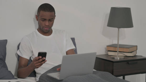 istock African Man with Laptop using Smartphone, Sitting in Bed 1421777515