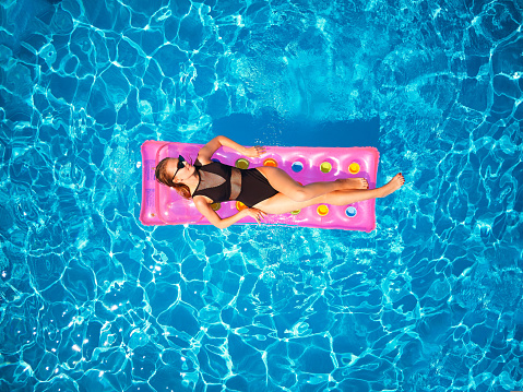 Fit pretty girl in bikini chilling on inflatable pink mattress in swimming pool. Slim hot woman in swimwear tanning. Female relaxing on a float in blue water at luxury resort. Aerial, view from above.