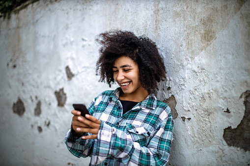 Beautiful young girl with afro hair and piercings, smiling, wearing a vintage plaid shirt lean against the wall, texting in the alley in Tay Ho, Hanoi, Vietnam