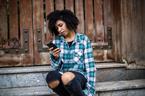 Beautiful young girl with afro hair and piercings, wearing a vintage plaid shirt, messaging, sitting on the stairs next to the old gate in Tay Ho, Hanoi, Vietnam