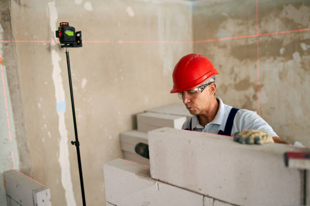 istock Bricklayer using rubber mallet hammer to tap and level concrete blocks wall. Contractor uses precise spirit bubble and laser level for brickwork. Worker constructs a wall in new apartment real estate. 1421780592