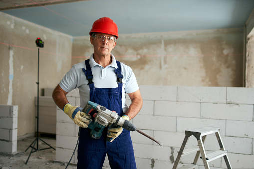 Professional construction worker in uniform standing with rotary hammer drill. Portrait of contractor in hardhat and overalls posing with a jackhammer near step ladder and masonry indoors.
