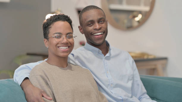 istock Portrait of Couple Smiling at Camera While Sitting on Sofa, Side Pose 1421781995