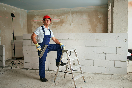 Professional construction worker in uniform standing with spatula on step ladder. Portrait of contractor in hardhat, overalls near masonry indoors. Handyman services for renovation with a copy space.