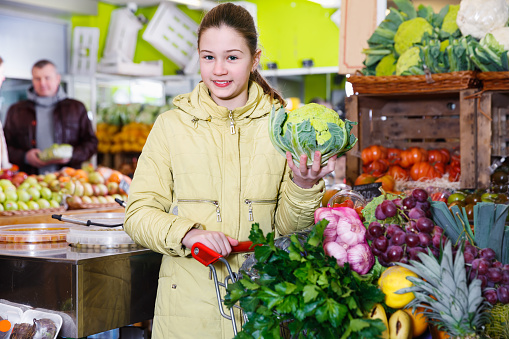 Smiling preteen girl choosing a ripe fruits and vegetables while shopping with parents in the greengrocery