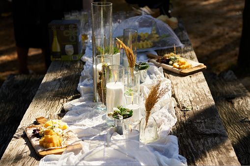 Wedding party banquet outdoors in forest. Dining table decorated in boho style with candles, white cloth, flowers, served with plates, tableware, dishes, meals, stemware and drinks. A holiday catering