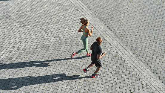 Top view of young black sports couple running on empty city area. Attractive slim curly girl and athletic man wearing sportswear. Concept of modern healthy lifestyle. Warm sunny day