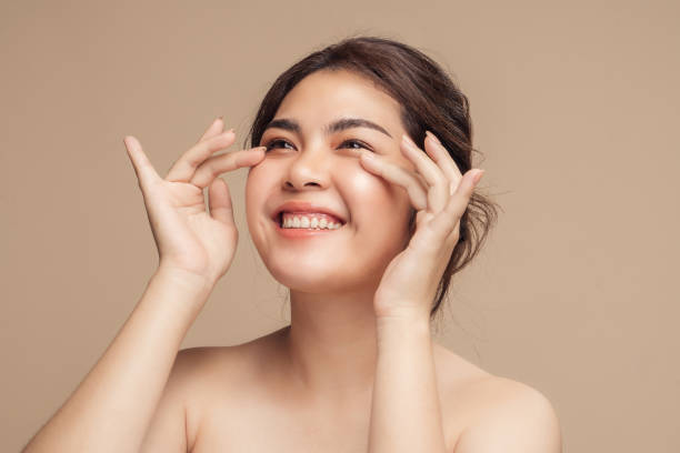 hermosa modelo femenina asiática sonriente con maquillaje natural que toca la piel hidratada brillante sobre fondo beige. - wrinkle treatment fotografías e imágenes de stock