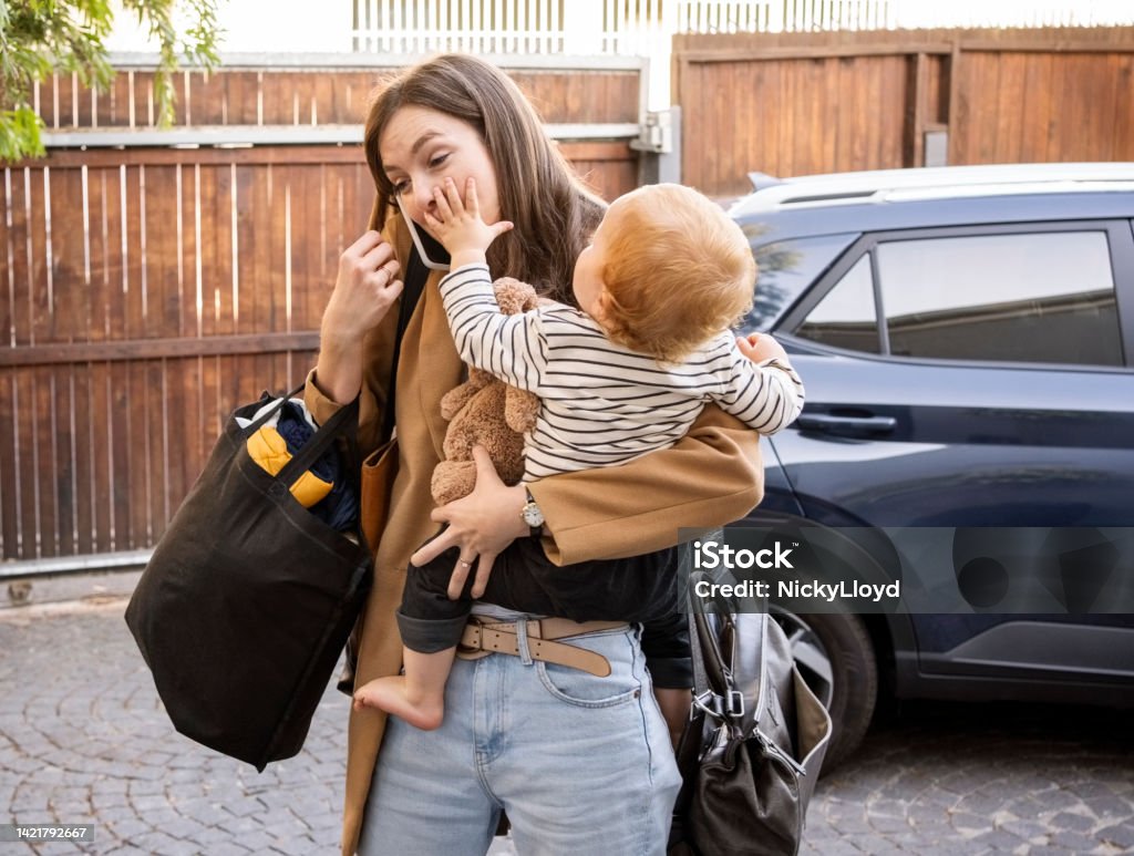 Woman carrying her son talking on mobile phone in driveway Woman carrying her son talking on mobile phone while walking in driveway with car parked at back Mother Stock Photo