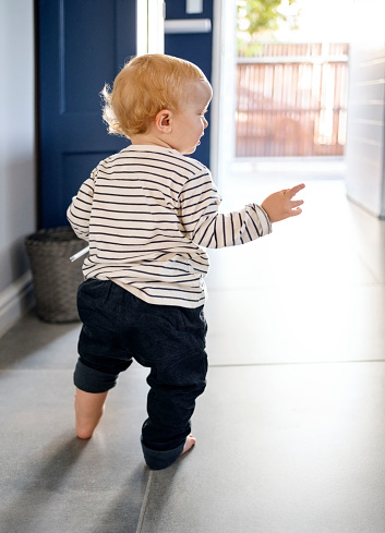 Rear view of cute baby boy learning to walk making his first steps inside the home