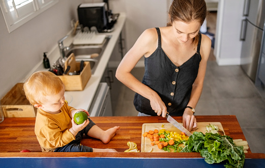 Mother cutting vegetables on chopping board in kitchen with son sitting on counter at home