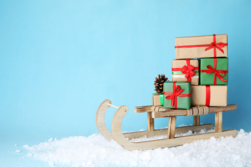 Wooden sleigh with Christmas gift boxes, pine cone and artificial snow on light blue background, space for text