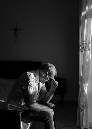 Monochrome of adult man sitting on bed against window with sunlight and shadow