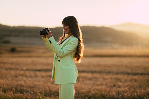 Fashionable young woman wearing a mint green pants suit taking photos with a camera on a sunny summer day in nature