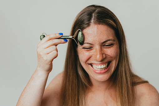 Young smiling woman with freckles using a jade roller on her face, studio shot in front of white background