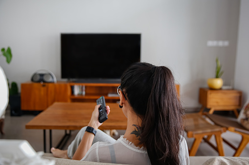 Rear view shot of mid adult indonesian woman turning on TV with remote control
