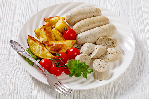 Weisswurst, bavarian white sausage of minced veal, pork back bacon, spices and parsley on white plate with roast potato, fresh tomatoes, horizontal view from above