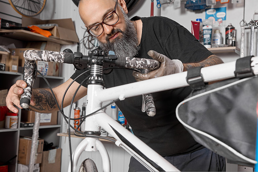 Bearded master in glasses adjusts position of handlebar on steering column of bicycle. Professional mechanic aligns the direction of the rudder relative to the steering column to the desired distance