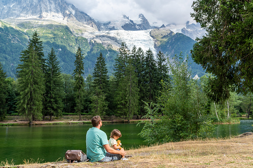 Father and his little daughter sitting on a lake shore looking at Mont Blanc slopes in Chamonix, France.