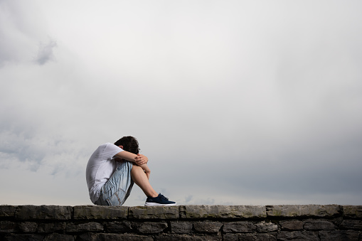 Sad teenager boy sitting under dramatic cloudy sky with copy space. Depression and mourning concept.