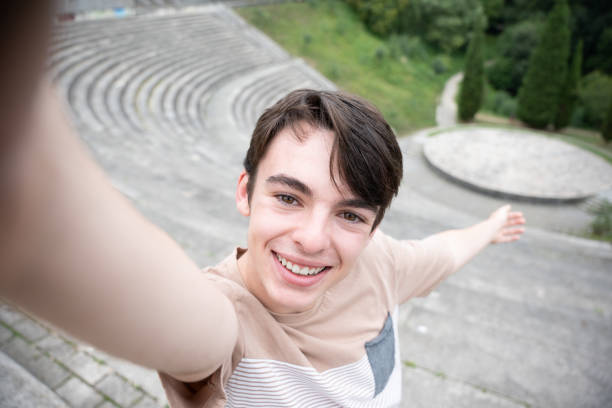Young teenager tourist boy taking a selfie in touristic destination. Happy adolescent looking and smiling at camera in amphitheatre
