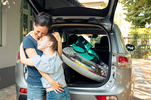 Mother and young kid hugging before travelling on holiday trip at seaside, loading luggage and suitcase in car trunk. Leaving on summer vacation with baggage and inflatable, leisure recreation.