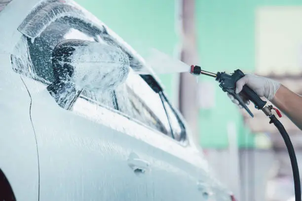 Man worker in garage using a high pressurised foam injector spraying on a dirty white car. Car washing and car care concept in rainy season. Manual labor spray a liquid soap on a dirty car.