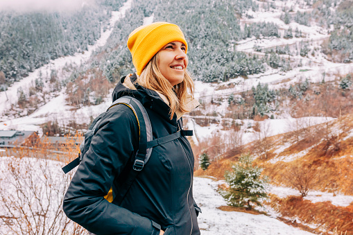 Winter hike on snow mountain young happy hiker woman climbing. Europe travel adventure trek in nature landscape. Young cheerful female wearing yellow hat, black jacket for cold weather and bag.