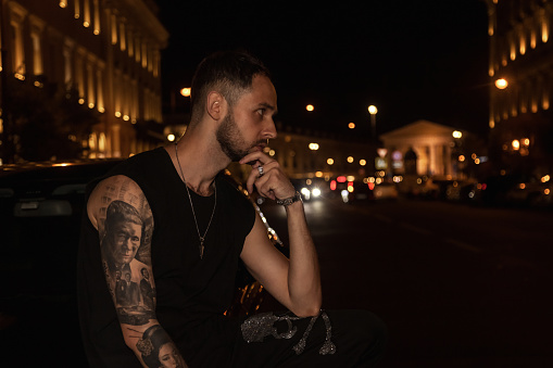 Young man sitting on car at night thoughtful, looking away, at urban background. Sad serious guy alone thinking about problems feeling lonely. Nightlife trendy youth atmosphere concept. Copy space