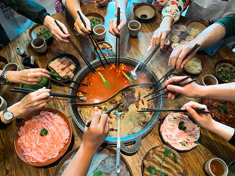 Many hands are holding chopsticks on two flavors hot pot with meat and tea on the dining table