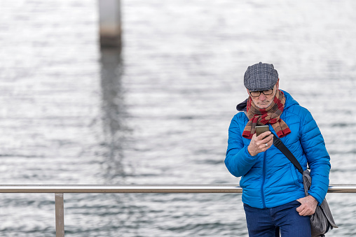 Senior man wearing eyeglasses leaning against railing on seafront while using smartphone