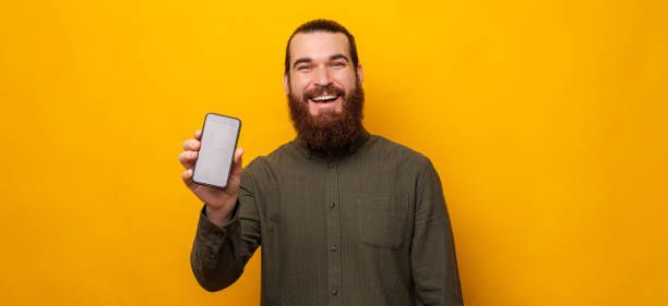istock Handsome bearded man shows phone screen to the camera over yellow background. 1421873815