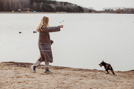 Happy enjoying blonde woman in warm clothes playing and having fun with cute puppy near river bank on cool sunny day, ducks swimming in water. Outdoors weekend activity, dogginess, water background.