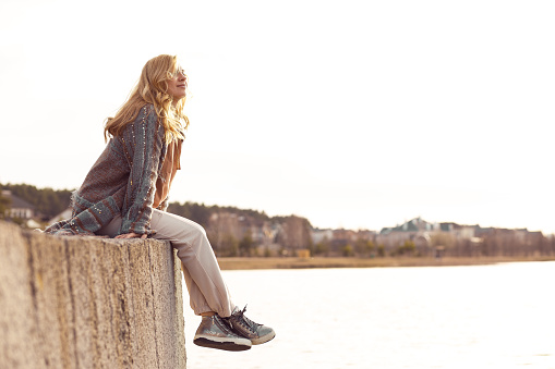 Happy smiling blonde woman sitting on river bank alone on cool sunny day, relaxing, meditating and enjoying peace and freedom. Outdoors weekend activity, scenic landscape, sky background.