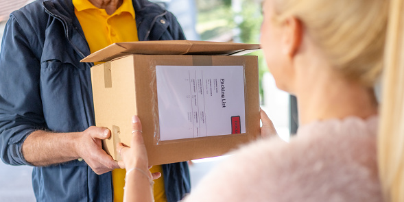Senior adult courier giving young female customer order, holding cardboard box, medium shot. Door to door delivery, postal worker and customer, home shipment service