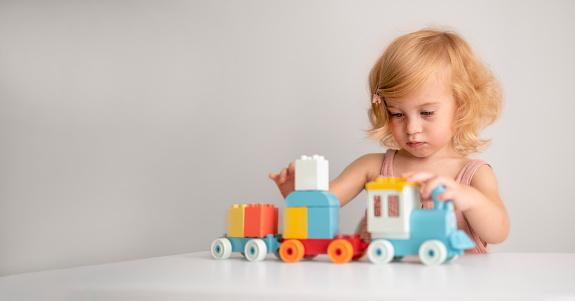 Pretty caucasian interested 1,2 year old with blond curly hair playing with colourful construction, erector set, toys making train.Nursery, preschool concept.Copy space.