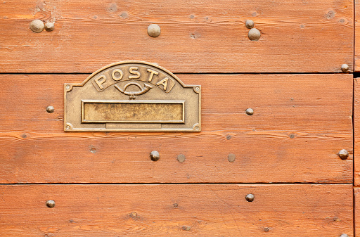 Metal maibox opening on the surface of a wooden exterior front door with the italian word for mail
