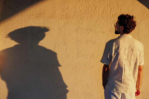 A portrait of a young stylish man standing in front of a wall during the sunset looking at his shadow on the wall.