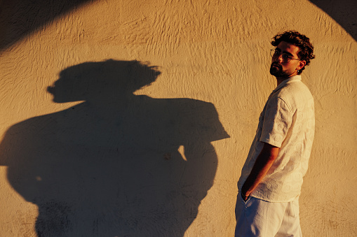 A young man is standing in front of a wall on a sunny day with his shadow being cast on the wall and looking over his shoulder at the camera.