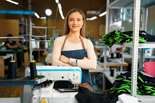 Portrait of a young smiling seamstress standing at working place and looking at camera