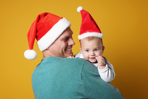 Happy father with cute baby wearing Santa hats on yellow background. Christmas season