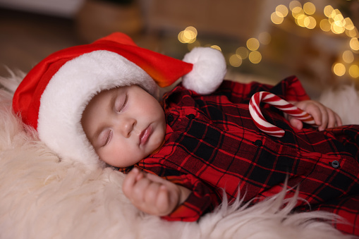 Cute baby in Santa hat with candy cane sleeping on soft faux fur indoors. Christmas season