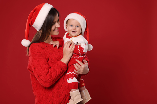 Happy mother with cute baby in Christmas outfits and Santa hats on red background, space for text