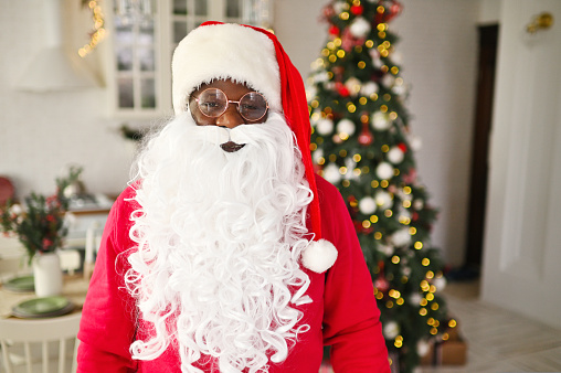 african american man in a santa costume smiles against the background of christmas decor and a fir tree.