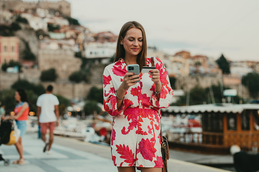 Young smiling paying with a credit card and a smartphone in the city street of the old town of Kavalla on a sunny summer day at the seaside