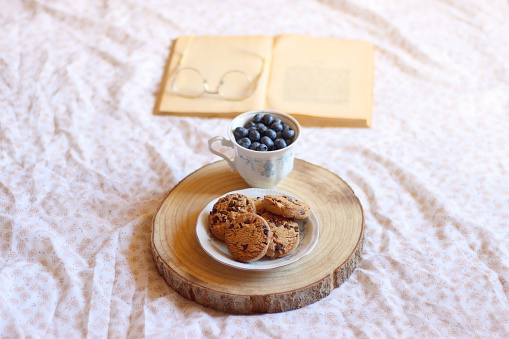 Vintage cup with fresh blueberries, plate of chocolate chip cookies, open book and reading glasses on a bed. Selective focus.