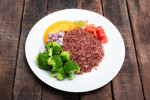Red rice in plate and vegetable broccoli tomato pumpkin on wood background close up, top view, healthy vegan food concept.