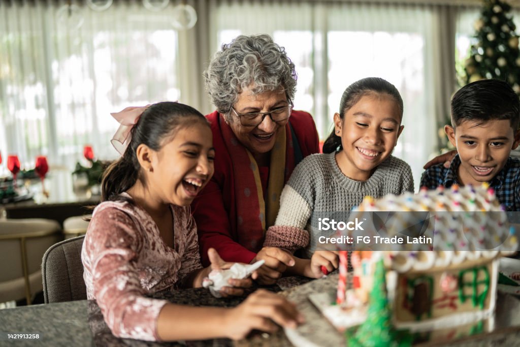 Grandchildren decorating a gingerbread house with grandmother for Christmas Grandchildren making a gingerbread house with grandmother at home Christmas Stock Photo