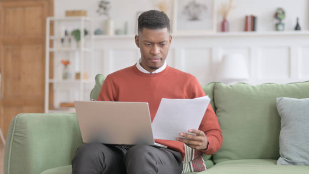 istock African Man with Laptop Reacting to Loss on Documents, Sofa 1421915344