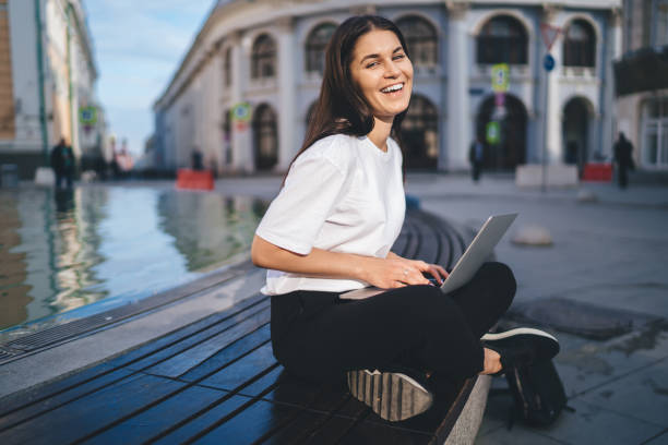istock Joyful hipster girl with modern netbook device laughing and posing while doing distance job, funny digital nomad with laptop computer smiling at camera catching free wi fi in touristic city 1421919926